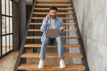 Poster - A man in a blue shirt and jeans sits on a set of wooden stairs, using a laptop and speaking on a phone. The stairs are made of wood and have a black metal railing