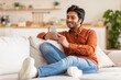 © Prostock-studio - A young Arab man in a casual orange shirt and blue jeans is sitting on a white couch and using his smartphone. He is looking at the phone screen and smiling. The room is a modern living room