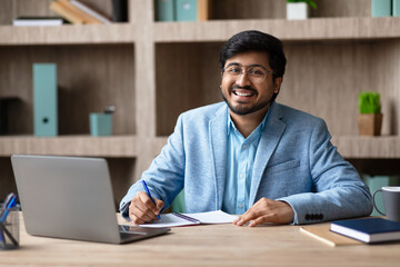 Aufkleber - A smiling Indian man with a beard and glasses sits at a desk in a modern office, working on a notebook while looking directly at the camera. He is wearing a blue suit jacket and a light blue shirt.