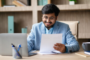 Poster - Indian businessman wearing glasses and a blue blazer sits at his desk, smiling as he reads documents. There is a laptop and a cup of coffee on the desk.