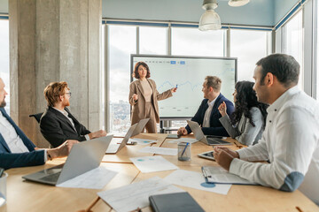 A businesswoman stands in front of a large screen and points to a graph as she presents data to a team of colleagues seated around a table in a modern office.