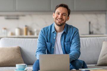 Poster - A man is sitting on a couch in his living room, smiling and looking at the camera while using his laptop. There is a cup of coffee on a table in front of him.