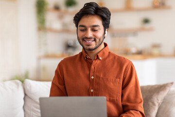 Wall Mural - Arab man sits on a couch in his home, smiling while looking down at his open laptop. He is wearing earbuds and an orange button-down shirt.