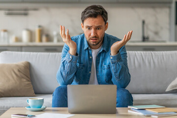 a man sits on a couch in his living room, looking confused as he stares at his laptop. his arms are 