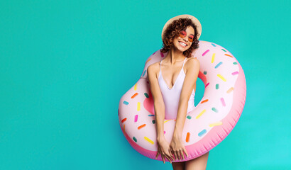 Wall Mural - A smiling black woman is standing in front of a teal backdrop, holding a pink donut-shaped inflatable pool float in front of her. She is wearing a white swimsuit, sunglasses, and a straw hat.