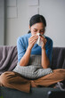 © amnaj - Young woman is blowing her nose with a tissue while sitting on a sofa at home, feeling unwell