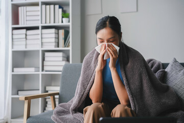 Young woman sitting on a sofa, wrapped in a blanket, and blowing her nose in a tissue