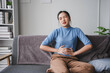 © amnaj - Young woman is sitting on her sofa, hunched over in pain and holding her stomach