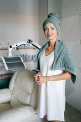 Wall Mural - Vertical portrait of attractive female with blonde hair in spa towel standing posing looking at camera after washing hair in beauty salon with casual atmosphere. Beauty and hair styling concept.