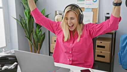 Sticker - Blonde woman celebrating with arms raised in a modern office wearing headphones