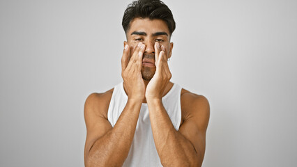 A young hispanic man applies cream on his face against an isolated white background.