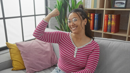 Poster - A smiling young hispanic woman in a striped shirt, wearing glasses, poses casually indoors, exuding confidence and beauty.