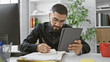 © Krakenimages.com - A focused man with a beard writing in a notebook and checking a tablet in a modern office setting