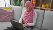 © Krakenimages.com - A young muslim woman in a pink hijab focused on her laptop while sitting on a gray sofa in a cozy living room.