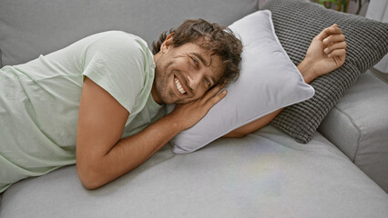 Poster - Handsome bearded hispanic man with green eyes lying on a sofa, smiling joyfully in a cozy indoor home setting.