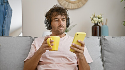 Canvas Print - Handsome hispanic man relaxing with coffee and smartphone on couch in cozy home interior