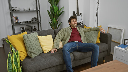 Wall Mural - A relaxed young hispanic man lounges casually on a gray sofa adorned with bright yellow pillows in a well-decorated living room.