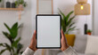 © Jane - Person holding black tablet iPad Pro vertical with white blank screen, mockup, sitting at a modern desk with a desk lamp and a green plant in the background