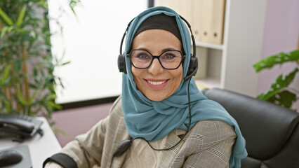 Sticker - A smiling mature woman wearing a hijab and headset sits in an office, representing professional customer support.