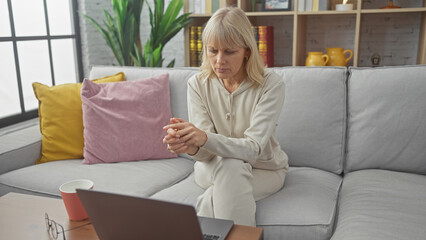 A pensive blonde woman in casual attire sits on a grey sofa indoors, with a laptop and mug nearby.