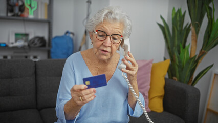 Concerned elderly woman holding credit card and telephone in her apartment living room.