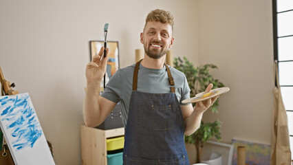 Smiling caucasian man with a beard, blue eyes, and an apron holding a brush and palette in an art studio.