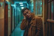 © Mandeep - Young man in brown jacket, standing in hallway with hand on ear, looking to side.