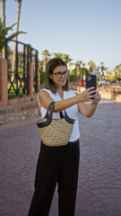 Sticker - Young woman taking a selfie in an outdoor setting with palm trees and paved ground, holding a woven handbag and wearing glasses