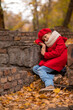 © Михаил Решетников - Sad caucasian girl in a red coat and beret sits on a brick wall on a walk in autumn.