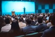 © MING - Photo of a college lecture with speakers on stage and an audience in the foreground, captured from behind against a blue colored background, with blurred people in focus.