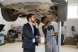 © sofiko14 - Mechanic in working overalls explaining car inspection details to business professional in auto repair shop. Businessman observing vehicle maintenance on lift with thoughtful expression.