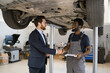 © sofiko14 - Professional mechanic and client shaking hands under raised car in auto repair shop. Smiling mechanic with clipboard in conversation with suited businessman.