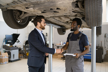 Professional mechanic and client shaking hands under raised car in auto repair shop. Smiling mechanic with clipboard in conversation with suited businessman.