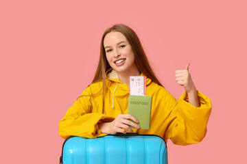 Poster - Smiling young woman in raincoat with passport and ticket showing thumb-up on pink background. Travel concept