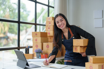 Small business entrepreneur, SME, young Asian woman Freelancer working from home standing holding a box Shipping information on  Laptops online, marketing, packaging, SME, ecommerce concepts.