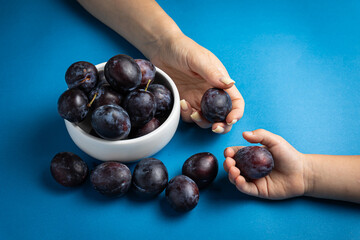 A child's hand reaches for a bowl of plums on a blue background