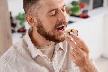 Wall Mural - Young man eating tasty muffin in kitchen, closeup