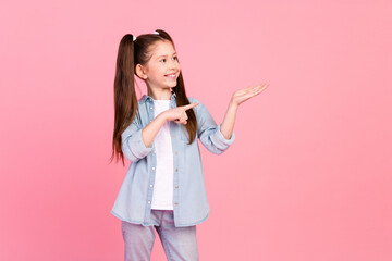 Poster - Photo of positive girl with ponytails dressed denim shirt directing look at object on palm empty space isolated on pink color background