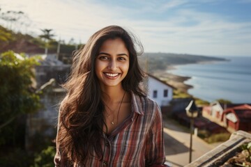 Poster - Portrait of a grinning indian woman in her 20s dressed in a relaxed flannel shirt in front of picturesque seaside village