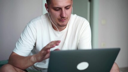 Young happy man talking on video webinar using laptop on bed at home. European programmer at an interview, talking with the team online. Remote remote online work concept.