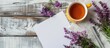 © HN Works - White wooden background with a blank sheet of paper pen tea filled yellow mug stationery and purple flowers Features space for text alongside a copy space image