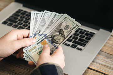 Paper dollar bills in the hands of a man against the background of a laptop, close-up, online earnings, a trader makes money on the Internet.