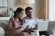 © Raool - Serious indian couple paying bills online, sitting on couch, using laptop, concentrated husband and wife checking papers and banking on Internet, copy, Generative AI
