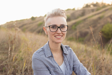 Stylish smiling woman portrait with short haircut hairstyle in glasses wearing blue shirt in nature outdoors on sunset near green hills