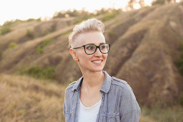 Stylish smiling woman with short haircut hairstyle in glasses wearing blue shirt in nature outdoors on sunset near green hills