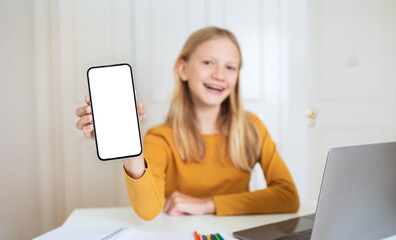 Fototapete - A teen girl with blonde hair sits at a desk with a laptop in front of her. She is holding up a smartphone with a blank white screen toward the camera and smiling.