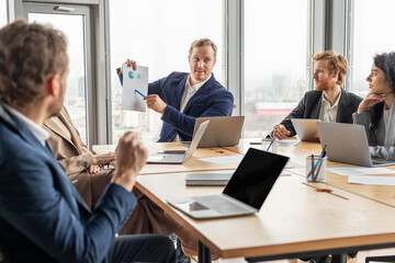 Aufkleber - A group of business professionals are gathered around a table in an office. One man is presenting data from a document to the group while the other colleagues listen attentively, engage in discussion.