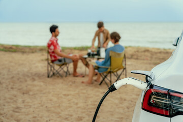 Wall Mural - Alternative family vacation trip traveling by the beach with electric car recharging battery from EV charging station with blurred family enjoying the seascape background. Perpetual