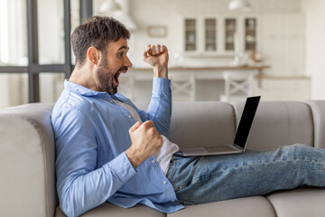 Poster - A man sits on a gray couch, his body turned toward the left and his face expressing excitement. He is wearing a blue button-down shirt and blue jeans and has a laptop computer on his lap.