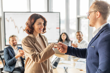 Aufkleber - A businesswoman in a beige blazer shakes hands with a male colleague in a blue suit, smiling broadly. The woman is standing, her colleague is seated, and they are both in a modern office setting